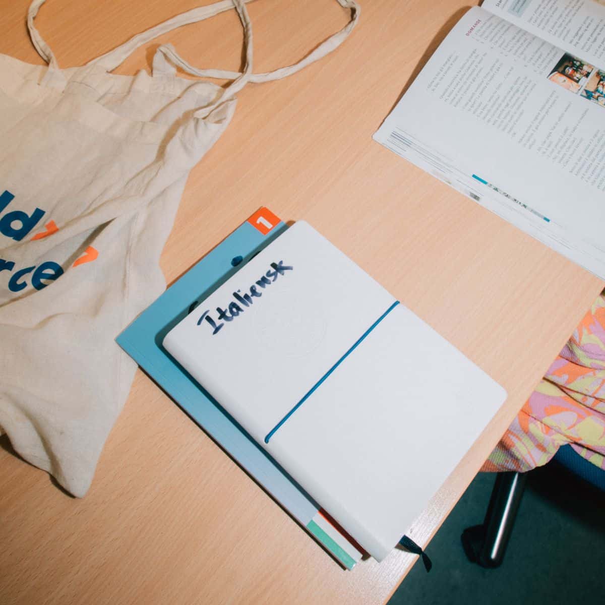 Noteshæfte med teksten “Italiensk” på skrivebord omgivet af undervisningsmaterialer – klar til sprogundervisning i italiensk.