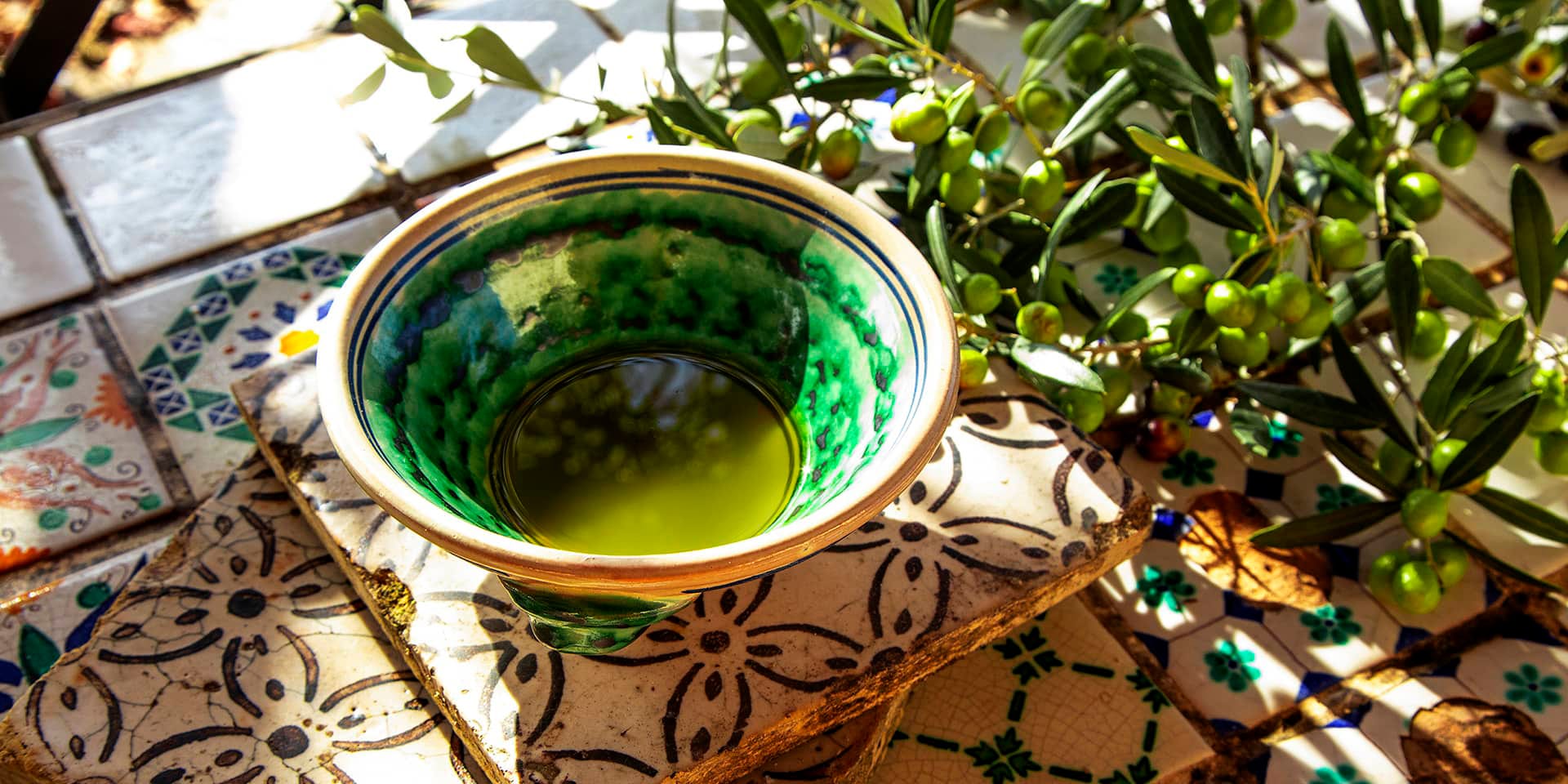 Olivenolie i en mønstret skål og olivengrene. På denne aften inviterer FOF Aarhus til smagning og foredrag om olivenolie.