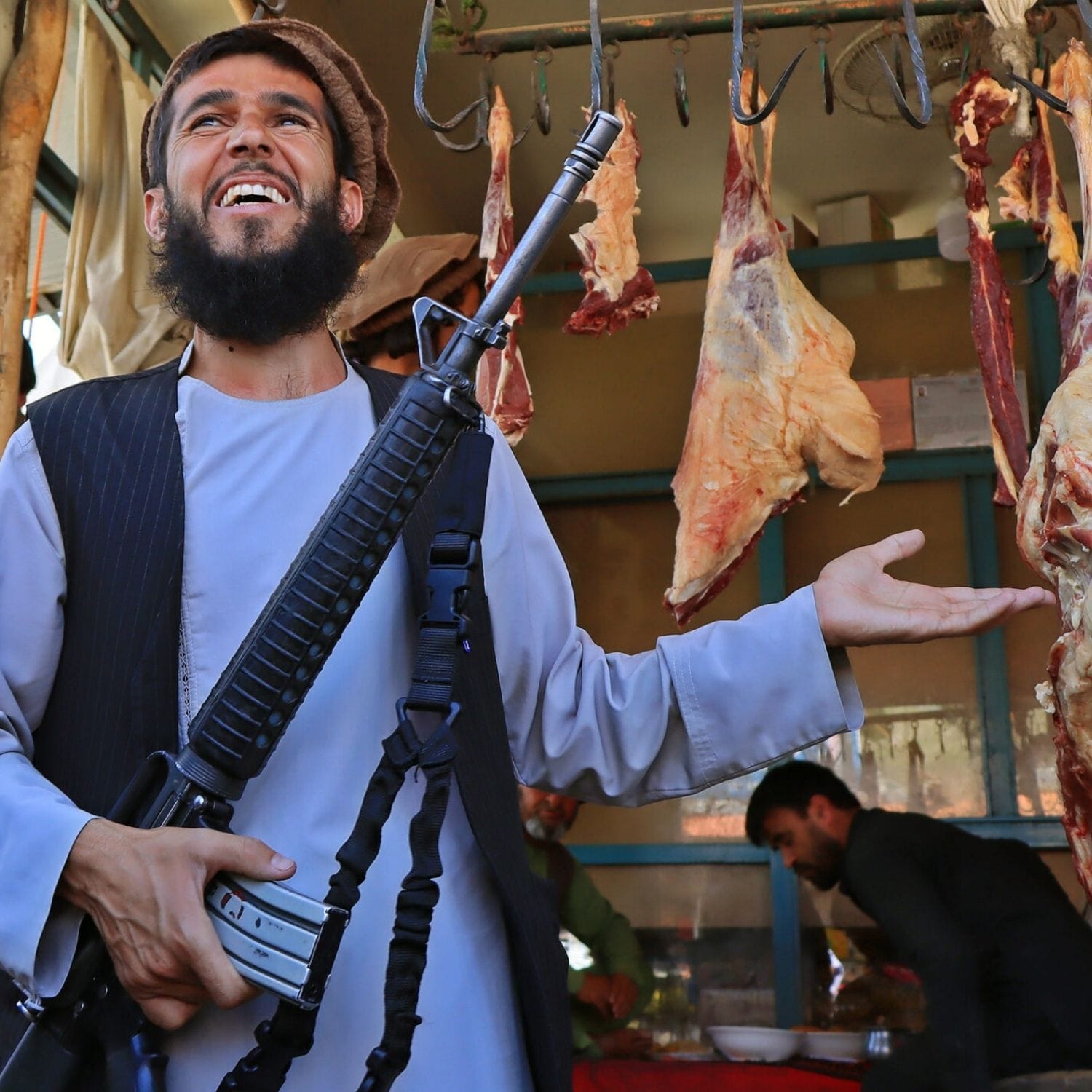 Taliban soldat der smiler med et maskingevær foran en butik med slagtet kød der hænger til tørre