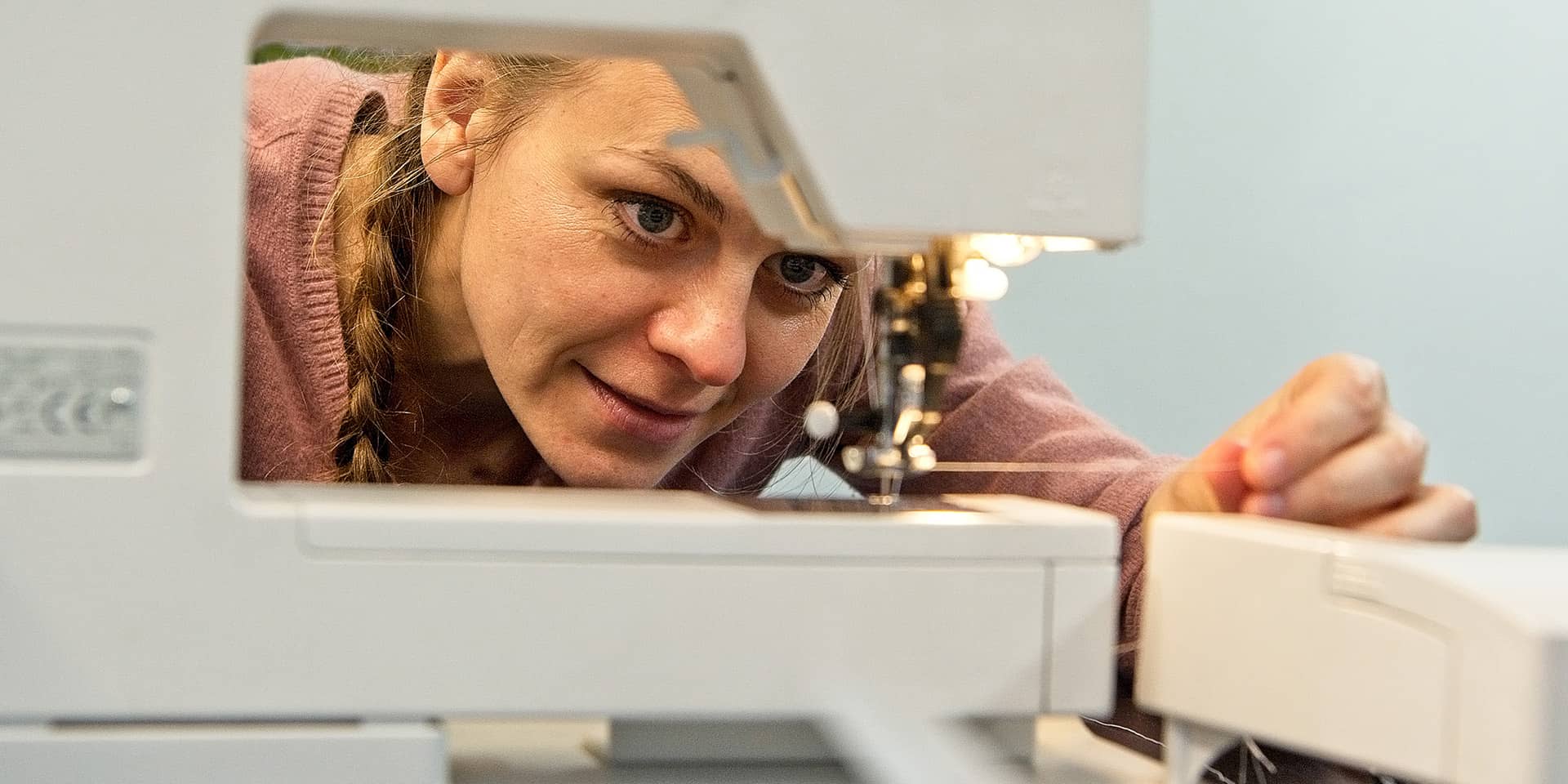 En kvinde, der er ved at sy. Hun sidder ved en symaskine og kigger på tråden og nålen i symaskinen. Til sykursus: lær at sy i FOF Aarhus