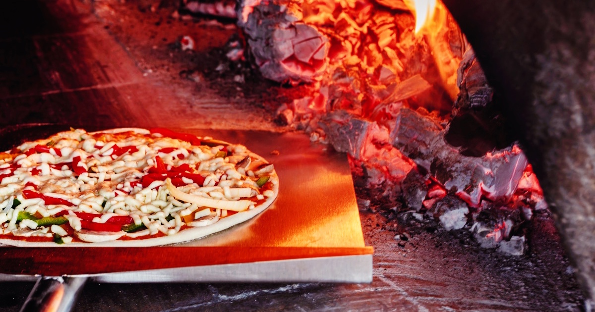 Pizza bliver sat ind i en brændefyret stenovn med åben ild og glødende kul.