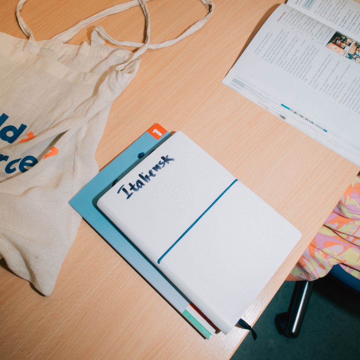 Noteshæfte med teksten “Italiensk” på skrivebord omgivet af undervisningsmaterialer – klar til sprogundervisning i italiensk.