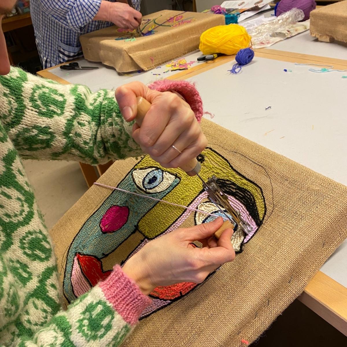 Kursister tufter små billedtæpper med Danella-nål ved et arbejdsbord.