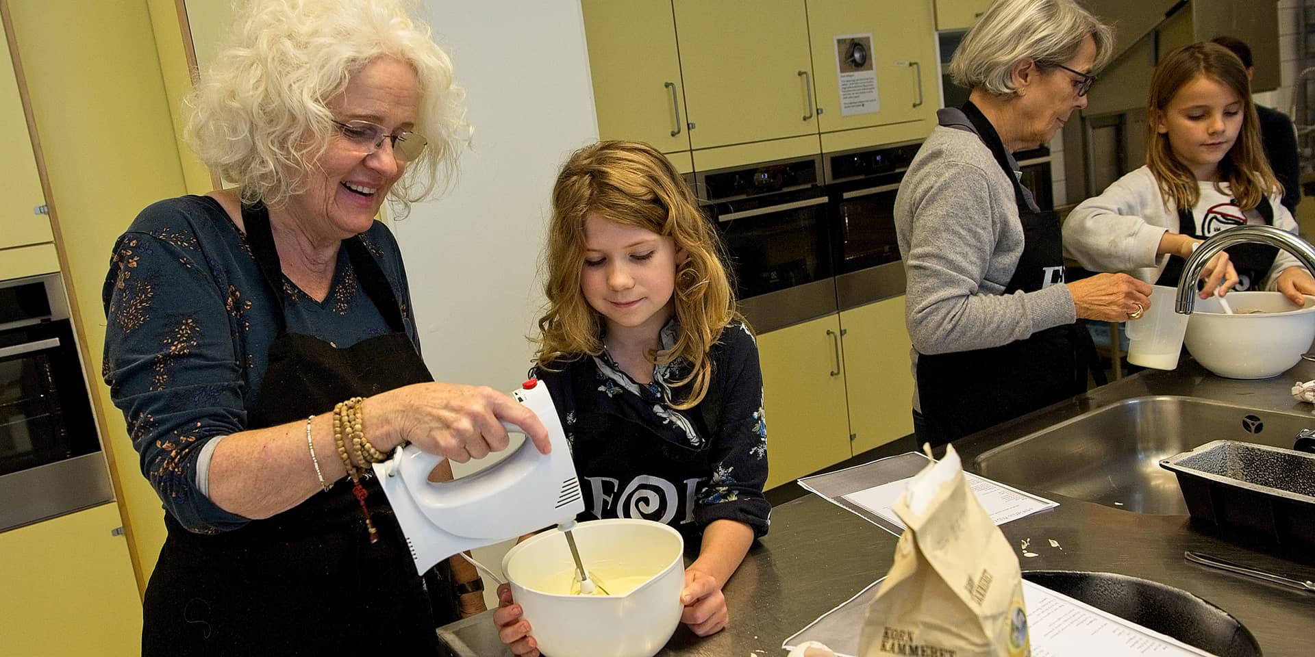 En kvinde, der pisker med en el-pisker, og en pige, der kigger på. Yderst i billedet kan man se en anden kvinde. Fra et madlavningskursus i FOF Aarhus for børn og voksne, hvor de laver flødeboller